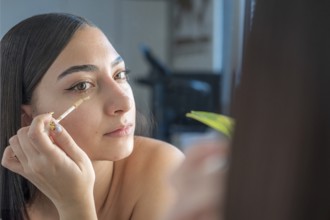A woman applies concealer under her eyes using a makeup wand, focusing on her reflection in the