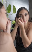 A woman in a black outfit applies makeup in front of a mirror. She holds a makeup brush near her