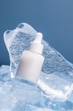 A serum bottle with a dropper rests on textured melting ice cubes against a blue background,