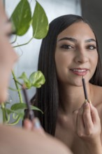 A woman applies lip liner while smiling at her reflection in a mirror in makeup session. A green