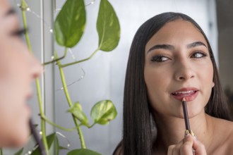 A woman applies lip liner while looking in a mirror, with lush green leaves in the background. The