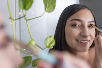 A woman is seen smiling while applying eye makeup in front of a mirror for the makeup session. A