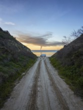 A lone camper van is parked on a dirt path leading to a tranquil Greek seaside. The sky is painted