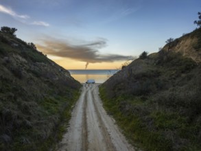 A narrow road flanked by green hills leads to a tranquil beach in Greece. A van is parked near the