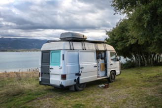 Camper van parked beside a scenic lake in Greece, surrounded by lush greenery and mountains. Cloudy