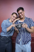 Charming gay couple smiling while reviewing photos on a vintage camera against a red wall. Casual