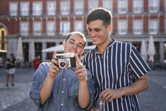 Happy gay couple reviewing photos on a vintage camera in Madrid's Plaza Mayor. Casual fashion,