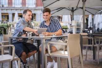 Gay couple sitting at an outdoor cafe in Plaza Mayor, Madrid, smiling while using a tablet and