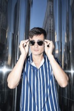 Fashionable young man in a striped shirt adjusting his sunglasses while posing against a reflective