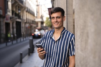 Young man in a striped shirt holding a smartphone and smiling on a city street. Relaxed and