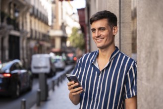 Stylish young man holding a smartphone and smiling on a vibrant city street. Dressed in a striped