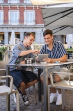 Gay couple sitting at an outdoor cafe in Plaza Mayor, Madrid, smiling while using a tablet and
