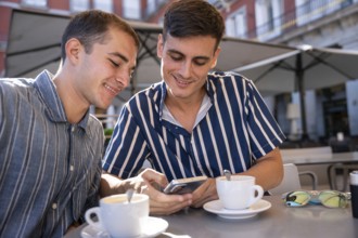Smiling gay couple enjoying coffee while browsing a smartphone at an outdoor cafe in Plaza Mayor,