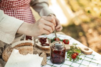 A person prepares canapes using fresh bread, sun-dried tomatoes, and herbs on a picnic table. The