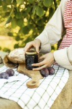 Hands preparing canapes outdoors on a wooden board. Fresh plums, homemade jam, and artisan bread on