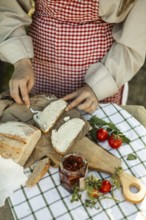 A person wearing a red checkered apron spreads cream on bread slices, surrounded by fresh tomatoes