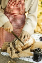 A person slices a loaf of fresh bread on a wooden board, wearing a red check apron. A jar of spread