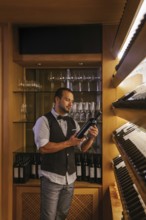 A professional sommelier stands in a cocktail bar, carefully examining a wine bottle. The shelves