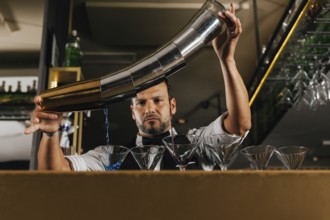 A professional bartender elegantly pours a vibrant blue cocktail into lined-up glasses at a chic