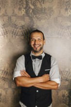 A professional bartender stands confidently with folded arms, wearing a stylish vest and bow tie.