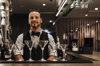 A professional bartender stands confidently behind a stylish bar adorned with martini glasses,