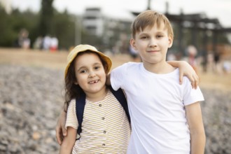 Two smiling siblings embrace while standing on a pebble beach. The young girl wears a sunhat, and