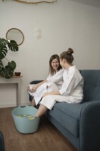 A mother and daughter engage in a self-care routine at home, wearing bathrobes and enjoying a foot