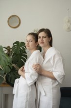A mother and daughter are enjoying a self-care day at home. They are wearing white bathrobes,