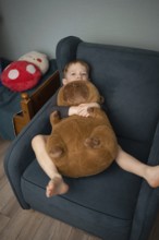 A child relaxes in a cozy chair, cuddling a large plush bear. The scene exudes comfort and warmth,