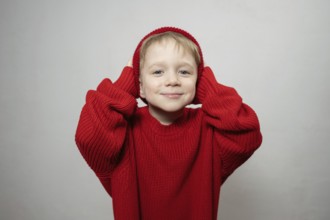 A cheerful young child wearing an oversized red sweater and hat, smiling playfully at the camera.