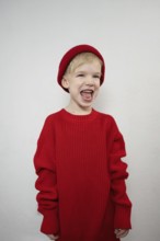 A cheerful child in a cozy red sweater and matching beanie laughs joyfully against a plain backdrop