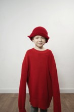 A child wears an oversized red sweater and matching hat, smiling playfully. The neutral background