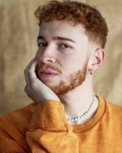A close up portrait of a stylish young man with red curly hair, wearing a pearl necklace and