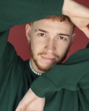 A man red hair wearing a green sweater poses with arms creatively framing their face against a red