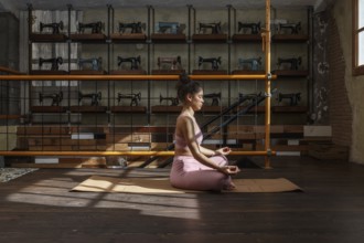 In a sunlit room filled with vintage sewing machines, a woman meditates on a yoga mat, practicing
