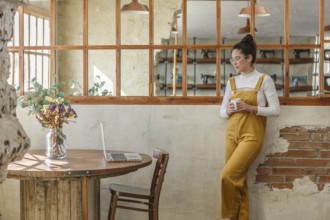 A young woman, holding a coffee cup, engages in telework at a rustic cafe table with her laptop