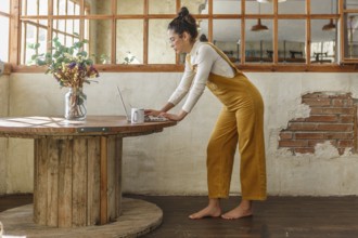 A young woman, dressed in yellow overalls, is focused on her laptop at a wooden circular table in a
