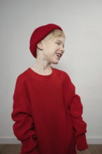 A joyful child wearing an oversized red sweater and matching beanie, smiling widely. The plain