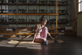 Woman in athletic attire practices yoga on a mat while working on a laptop. Sunlight filters