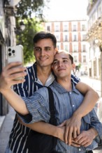 A gay couple embraces while taking a selfie on a picturesque street in Madrid. Sunlight filters
