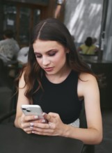 A woman in a black top intently uses her smartphone while sitting at a cozy cafe. The background