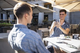 A gay couple enjoys coffee at an outdoor cafe in Madrid. One smiles, capturing the moment on his