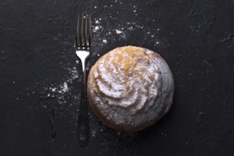 Top view of a single cream bun, generously sprinkled with icing sugar, rests next to a fork on a