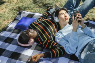 Two friends enjoy a sunny day, lying on a checkered blanket. One listens to music with headphones,