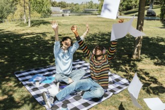 Two university students celebrate joyfully in a sunny park. Sitting on a picnic blanket, they toss