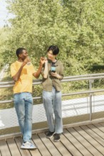 Two friends walking on a deck with lush green trees in the background, enjoying drinks, smiling,