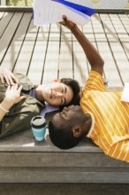 Two friends lying on a wooden deck. One is holding papers above them, while a coffee sits nearby.