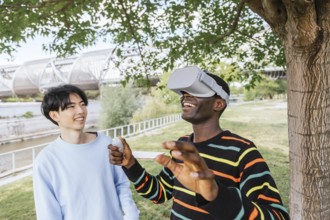 Two friends enjoy a VR experience outdoors. One wears a VR headset, immersed in a virtual world,