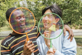 Smiling friends hold colorful badminton rackets over their faces with a shuttlecock. This image