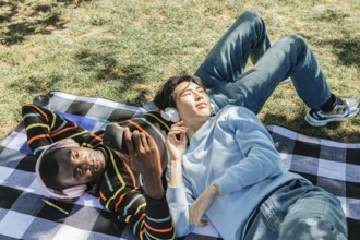 Two friends enjoy a sunny day in the park, lying on a checkered blanket. They are wearing